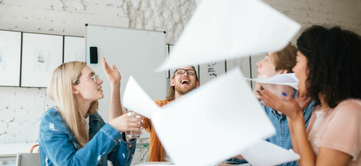 Emotional people happily throw up papers in office while working together. Group of young students joyfully studying with books on table in classroom.