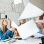 Portrait of emotional people happily throw up papers in office while working together. Group of young students joyfully studying with books on table in classroom.